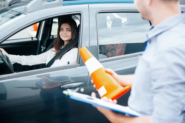 Attractive brunette woman on drivers seat with fastened seat belt smiles to male driving instructor with clipboard and orange traffic cone in his hands. Improving driving skills concept.