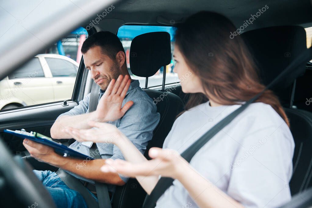 Young worried woman in white shirt and angry male driving instructor emotionally argue over results of driving test. Young man in grey shirt on passengers seat makes stop gesture with his hand.