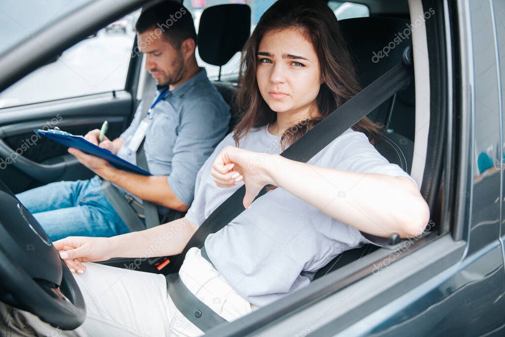 Sad female student fails her driving test and shows thumbs down. Attractive woman sits behind the wheel of a car and looks out the side window. Male instructor on front passenger seat takes notes.