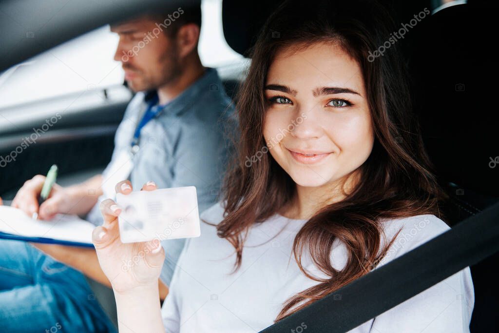 Attractive young woman showing drivers licence, looking at camera and smiling. Brunette woman sitting on drivers seat with fastened seat belt. Blurred male instructor on background writing something.