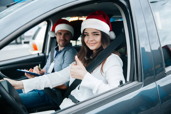 Young woman showing thumb up. Merry Christmas and Happy New Year Driving courses on winter holidays. Female student and instructor in santa hats smiling and looking into camera through car window.