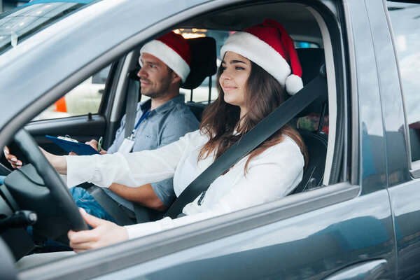 Driving school on christmas holidays. Young woman in red santa claus hat and white clothes holds the steering wheel and smiles. Male instructor with badge on his neck looks on the road. New Year.