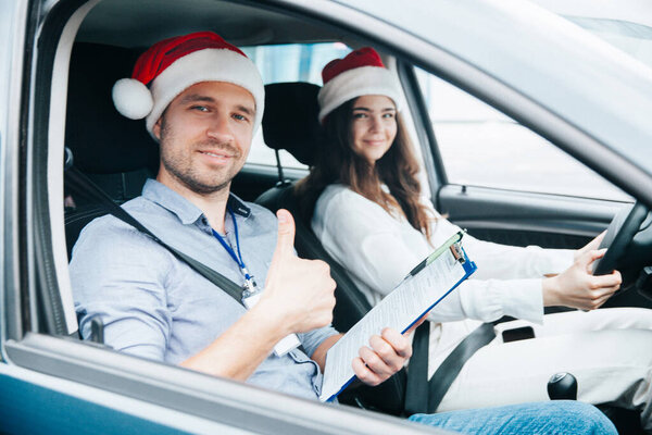 Young female driving student and male instructor in santa claus hats sit in a car together. Thumb up gesture. View through car window. Merry Christmas and Happy New Year. Driving courses concept.