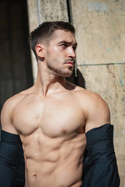 屋外で肩を脱いだシャツを着たハンサムな筋肉質の男 男は壁にもたれて立っている 強いセクシーなアスリート — ストック写真