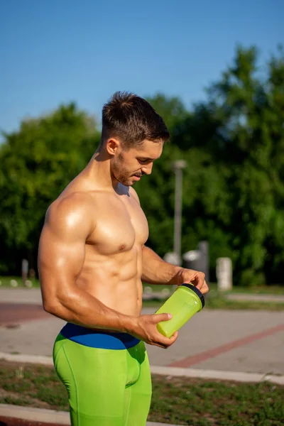 筋肉のボディビルダーの男が立って運動後に水を飲んでいます トレーニングの後に残っている恥知らずの若いスポーツマン — ストック写真