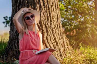 Orta yaşlı bir kadın bir ağacın altında oturmuş parkta kitap okuyor.