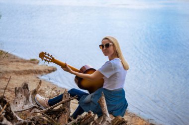 Beyaz tenli güzel sarışın kadın nehir kenarındaki kütükte oturup gitar çalıyor. Meditasyon, ekoloji, uyum, maneviyat, sağlıklı yaşam, barış konsepti. Arkadan bak.