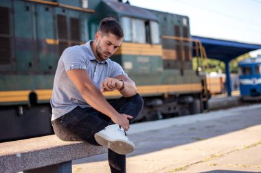 Genç kaslı bir adam tren istasyonunda oturmuş birini bekliyor ve saatine bakıyor.