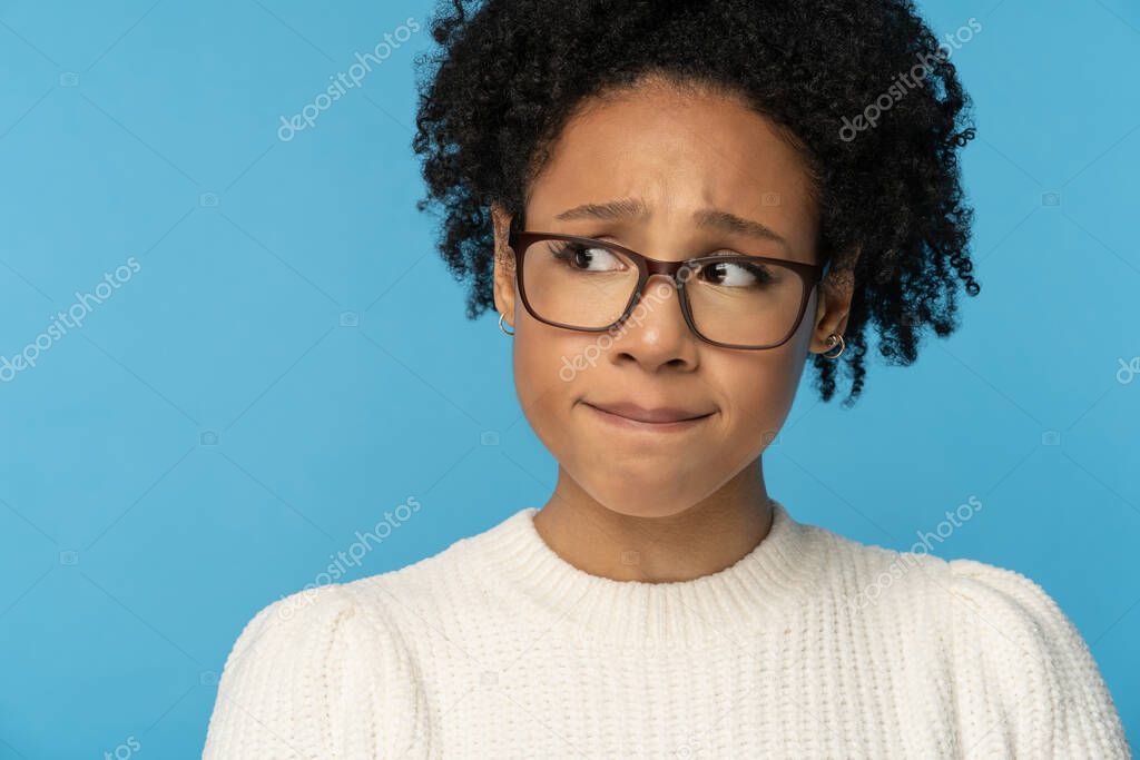 Close up studio portrait of shy awkward young Afro American woman wear glasses white sweater, biting lips feeling embarrassed, confused and nervous, looking aside, isolated on blue background.