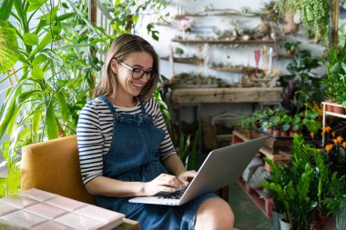 Woman gardener in glasses wear blue jeans overalls, sitting on chair in greenhouse, using laptop after work, communicates on internet with customer surrounded by plants. Home gardening, freelance. clipart