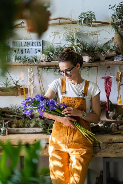 Surprised florist woman in glasses, wear orange overalls surrounded by collection of tillandsia air plants, charmed with pleasant gift, smiling, looking at a bouquet of irises flowers. Indoor garden