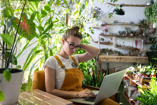 Woman gardener in glasses wear orange jeans overalls, sitting on chair in greenhouse, working on laptop on some project surrounded by plants. Home gardening, freelance.