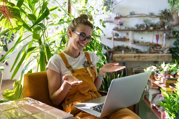 Woman gardener in glasses wear orange overalls, sitting on chair in green house, using laptop after work, smiling and speaking on video call surrounded by exotic plants.Home gardening, small business.