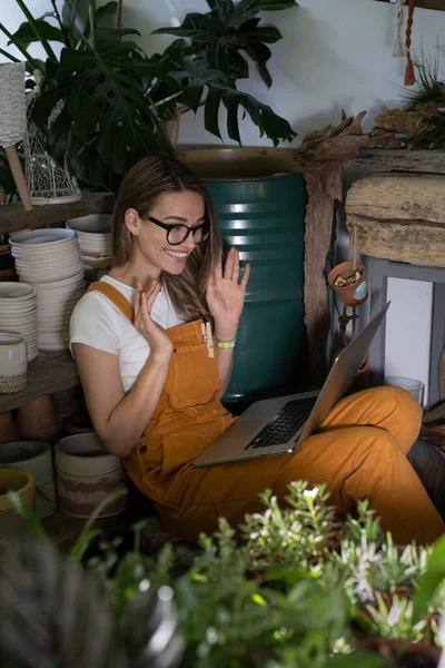 Cute woman florist in glasses wear orange overalls, sitting on the floor in flower shop, resting, using laptop after work, smiling and speaking on video call surrounded by plants. Work from home.