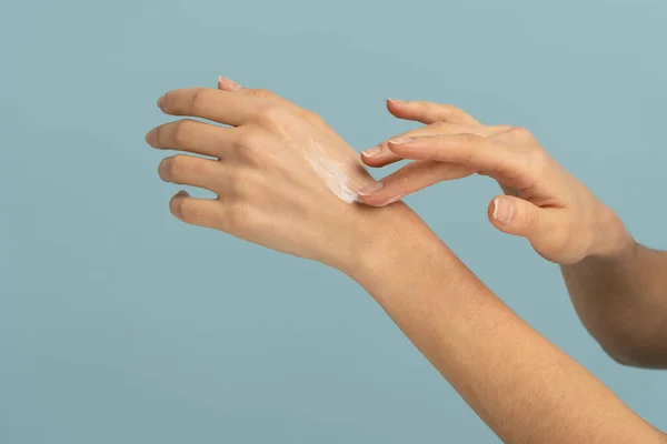 Mujer aplicando crema protectora sobre la piel seca de las manos en temporada fría, aislada sobre fondo azul estudio — Foto de Stock