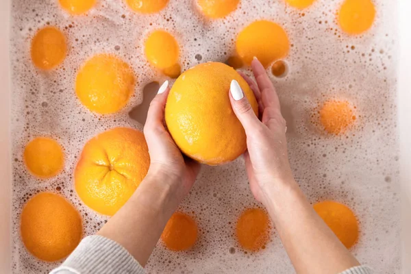 Top view of hands of woman washing ripe orange, grapefruit under faucet in the sink kitchen, soaking fruits in soapy water thoroughly washes after the store.