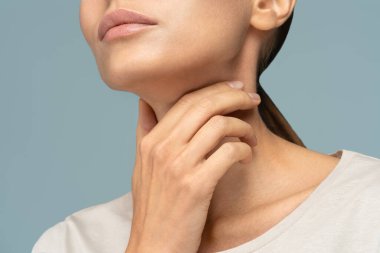 Closeup of sick woman having sore throat, tonsillitis, feeling sick, caught cold, suffering from painful swallowing, strong pain in throat, holding hand on her neck, isolated on studio blue background clipart
