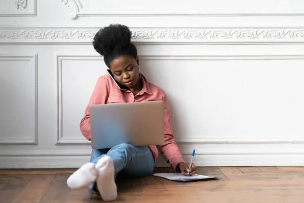 アフリカ系アメリカ人女性がノートを作成し 情報を明確にし 電話で話し ノートパソコンを使用して床に座っています モバイルデバイスによる出産女性従業員のコンサルティングは 自宅でリモートで動作します — ストック写真