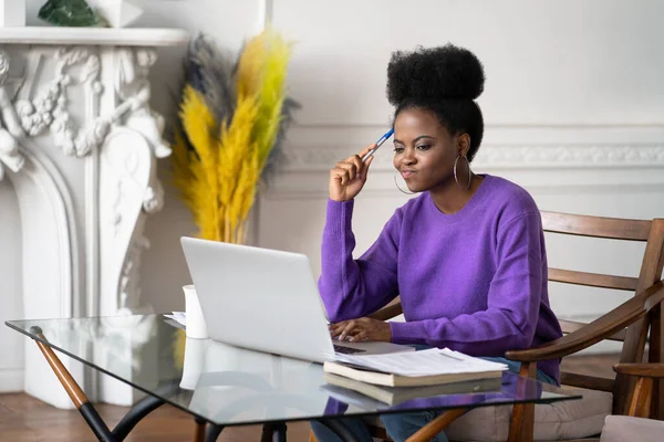 Afro-American millennial woman employee with displeased face working and looking at laptop, having operational software problems, annoyed with slow internet connection, doing homework, holding a pen