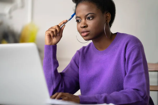 Afro-American millennial student woman with afro hairstyle browsing information on laptop, preparing for exam online, watching video course, holding a pen. Online education in zoom app, video training