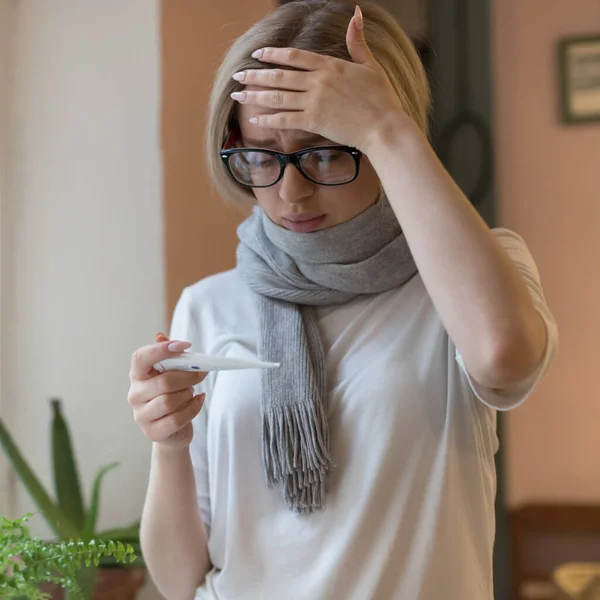 メガネとTシャツを着た若い女性が寒気のするスカーフに包まれ体温計を見て額に触れた ウイルスインフルエンザの季節 — ストック写真
