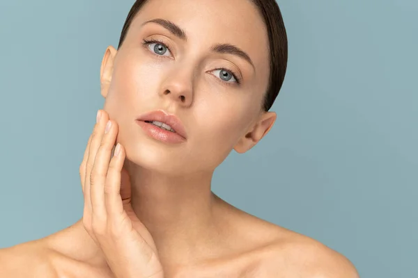 Studio portrait of young woman with natural makeup, combed hair, touching well-groomed pure skin on face, looks at camera, isolated on blue background. Beauty, facelift, anti-aging cosmetic procedure.