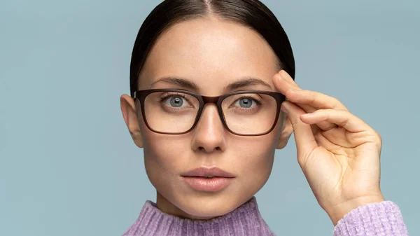 Self-assured serious business woman touches eyeglasses, looking at camera, isolated on studio blue background. Confident female with combed hair adjusts glasses, free space.