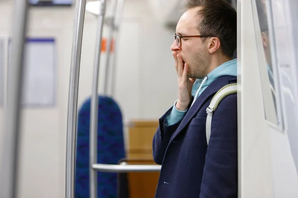 Tired man in eyeglasses in public transportation. Young exhausted male standing and yawning in subway, sleepy after work goes home, blurred background. Fatigue,sleep deprivation concept