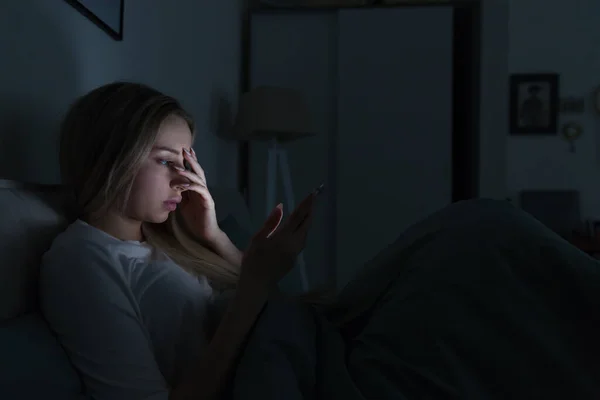 Young sleepy exhausted woman lying in bed using smartphone and touching her forehead, can not sleep. Insomnia, nomophobia, sleep disorder concept. Gadget addiction.