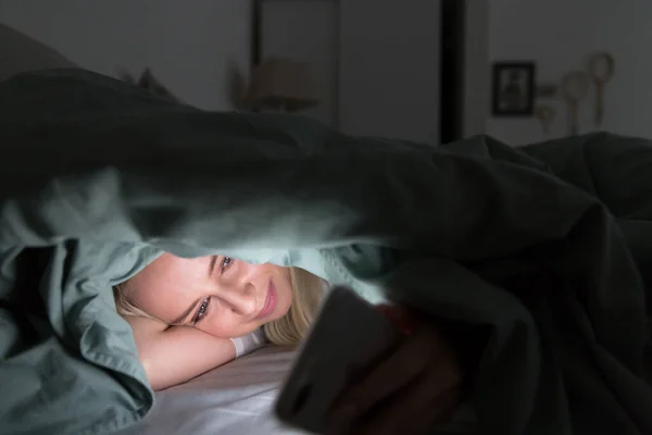Cheerful young woman lying in bed under the blanket using mobile phone at late night, can not sleep. Insomnia, nomophobia, internet addiction concept