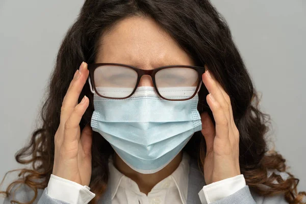 Femme d'affaires avec des lunettes brumeuses de souffle causé par le port d'un masque jetable sur le mur gris studio — Photo