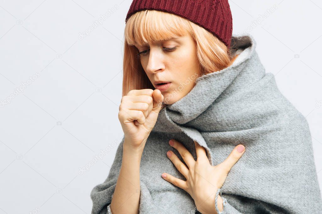 Cute sick young teen woman in red hat, wrapped in warm scarf coughing, closed eyes.Female feeling the first symptoms of illness.Bronchitis, upper respiratory infection concept.Moist cough, laryngitis