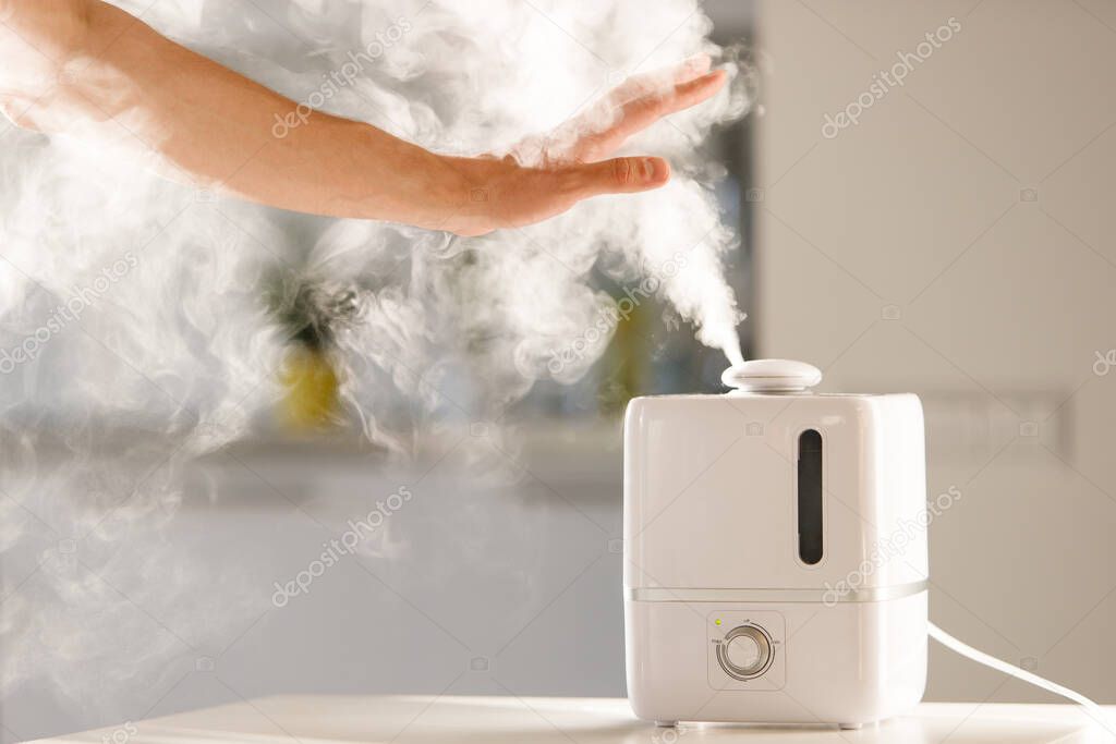 Man holds hand over steam aroma oil diffuser on the table at home, steam from the air humidifier, free space. Ultrasonic technology, increase in air humidity indoors, comfortable living conditions