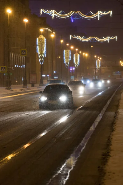 Bad winter weather. Cars driving on the roadway during a snowfall at night, road processed de-icing chemicals. Soft focus.