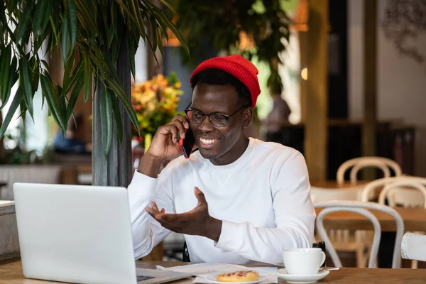 Feliz millennial hombre negro riendo, hablando con un amigo en el teléfono móvil, trabajo remoto en la cafetería. — Foto de Stock