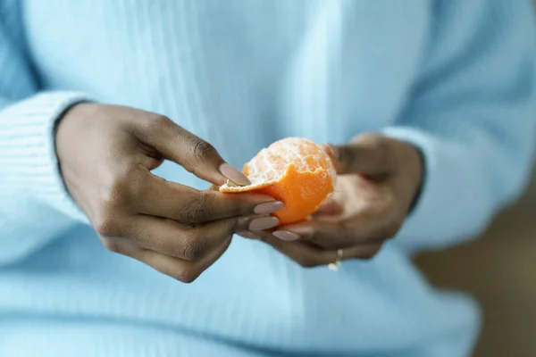 Afro nő meghámozza az érett mandarint, kék pulóvert visel, közelről. Téli gyümölcs, egészséges táplálkozás, Karácsony — Stock Fotó