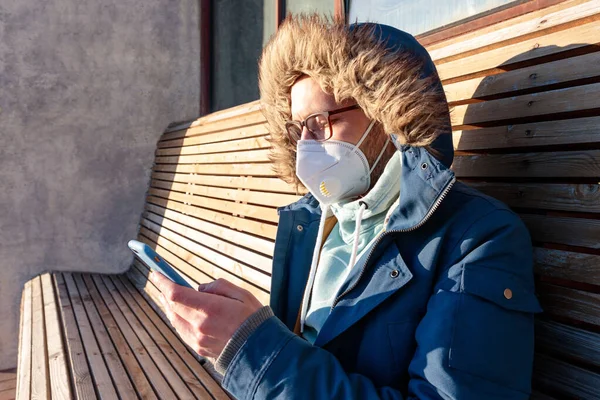 Sad man with hood sitting alone on bench in sunny weather, wearing facial mask against transmissible infectious diseases, coronavirus pandemic, using smartphone. Life in isolation and quarantine