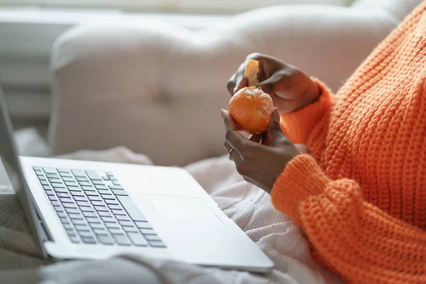 Afro mujer manos pelando mandarina dulce madura, usar suéter naranja, trabajando en el ordenador portátil en casa —  Fotos de Stock