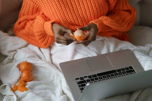 Afro mujer manos pelando mandarina dulce madura, usar suéter naranja, acostado en la cama debajo de la cuadros — Foto de Stock