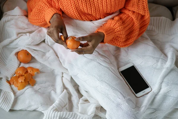 Tangan wanita Afro mengupas jeruk yang matang, mengenakan sweater oranye, berbaring di tempat tidur di bawah kotak-kotak — Stok Foto