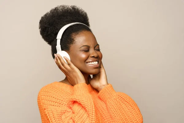 Souriant Afro femme en pull orange toucher écouteurs sans fil, écoute de la musique préférée isolé — Photo