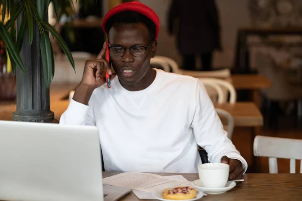 Černý hipster muž mluví na mobilním telefonu vzdáleně on-line práce na notebooku v kavárně drží šálek čaje — Stock fotografie