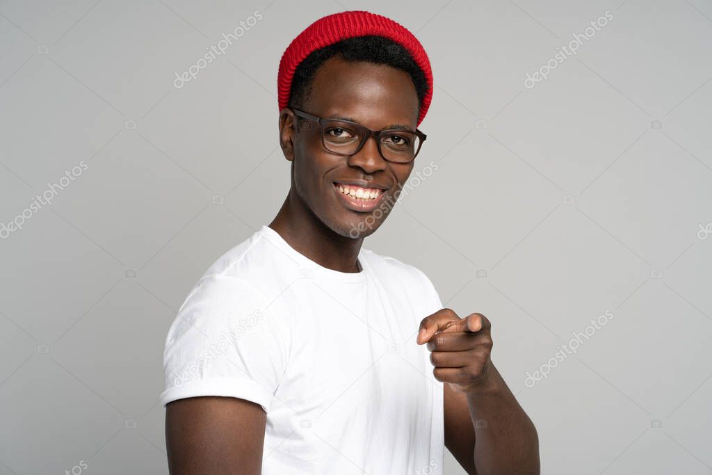 Cheerful positive Afro hipster man smiling broadly, pointing a finger at you, studio grey background