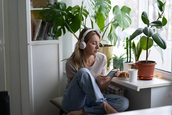 Frau, die Musik hört, trägt kabellose weiße Kopfhörer mit Handy, trinkt Tee zu Hause — Stockfoto