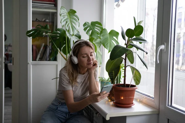 Frau, die Musik hört, trägt kabellose weiße Kopfhörer mit Handy, trinkt Tee zu Hause — Stockfoto