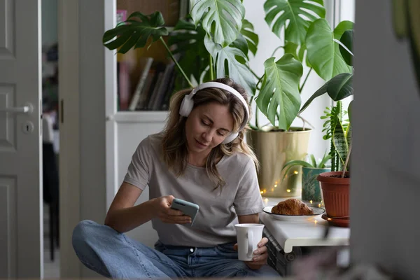 Kvinna som lyssnar på musik bär hörlurar, använder mobiltelefon chatta i sociala nätverk, dricka te — Stockfoto