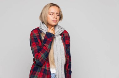 Upset young woman in checked shirt, wrapped in scarf having sore throat, holding hand on her neck. Throat pain, painful swallowing concept.  clipart