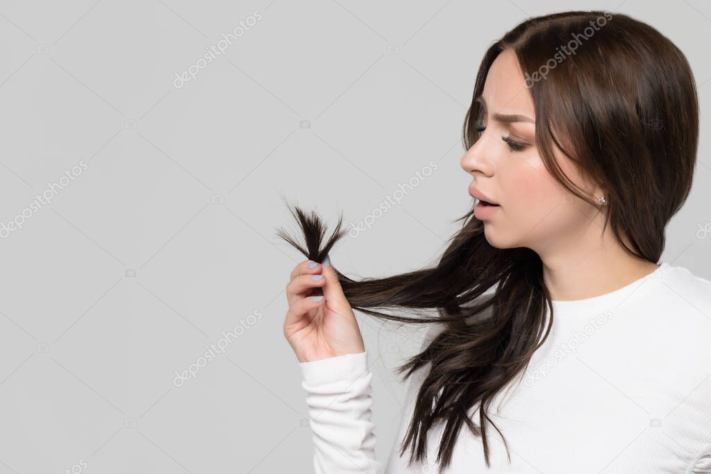 Closeup studio portrait of frustrated sad brunette woman holding and looking at split ends of her damaged hair. Separated dry ends, hair care problem