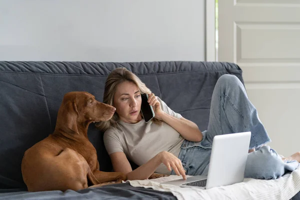 Woman lying on couch with Vizsla dog, talking on cellphone, remotely working on computer laptop — Stock Photo, Image