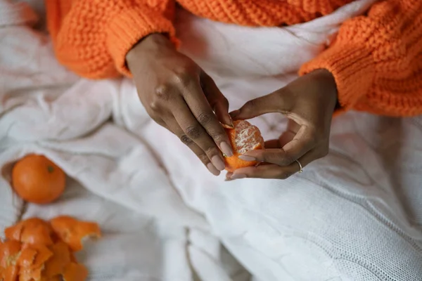 Афро женщина руки пилинг спелых сладкий мандарин, носить оранжевый свитер, лежа в постели под клеткой — стоковое фото